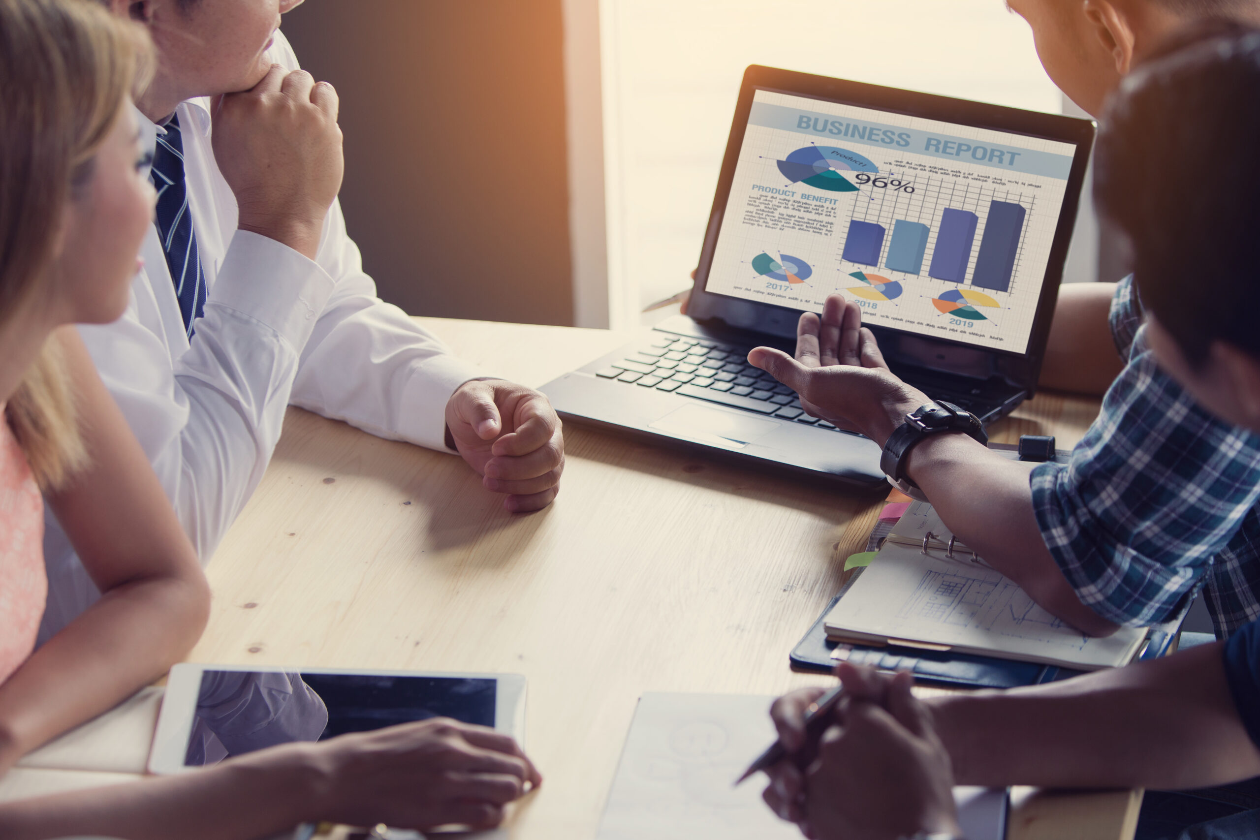 A group of professionals gathered around a table, discussing charts and graphs displayed on a laptop screen.