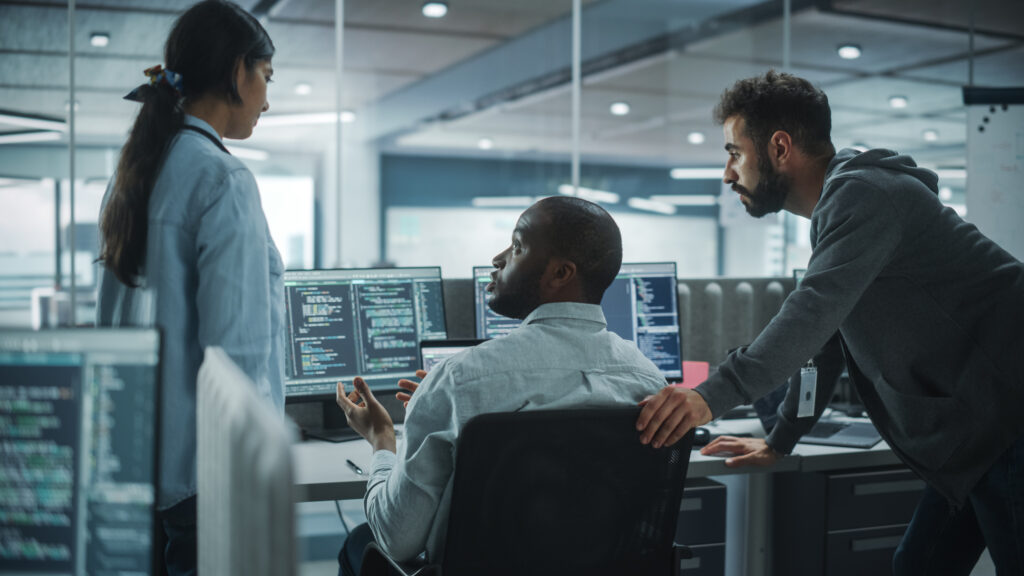 A team of three colleagues discussing code on multiple computer screens in a sleek, modern office space.