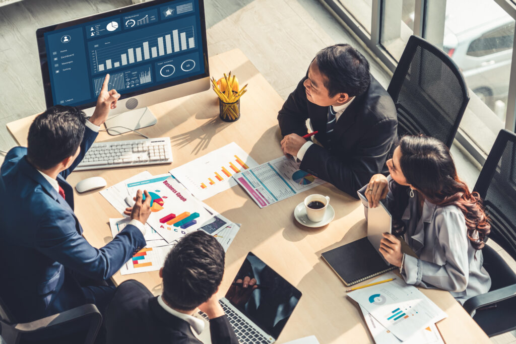 A group of professionals reviewing charts and graphs on a digital display during a meeting, with documents and laptops on the table.